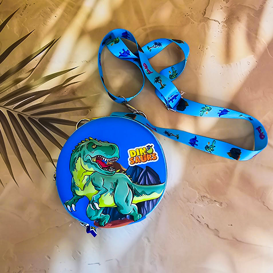 Two colorful, round kids’ sling pouches with attached lanyards are placed on a sandy beige background with a tropical leaf shadow. The left pouch is purple and features an adorable cartoon unicorn with a pink mane, large sparkling eyes, and the name "SHYAMA" written in white. It has a lilac lanyard decorated with candy prints. The right pouch is bright blue with a fierce green cartoon T-Rex dinosaur illustration labeled “DINOSAURS” in bold yellow text. Its lanyard is blue with black dinosaur silhouettes. Bo